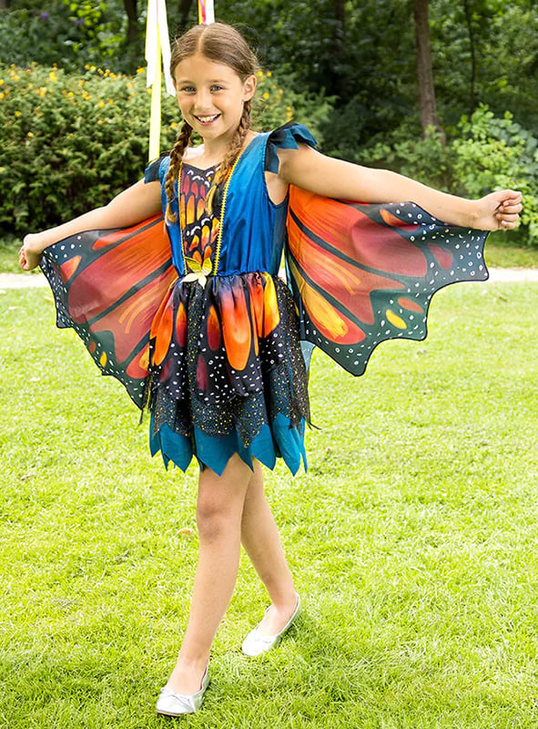 Ein junges Mädchen steht lächelnd draußen auf dem Rasen und trägt ein Kleid mit Schmetterlingsflügeln – ein farbenfrohes Kinderkostüm mit wunderschönen, weit ausgebreiteten Flügeln.