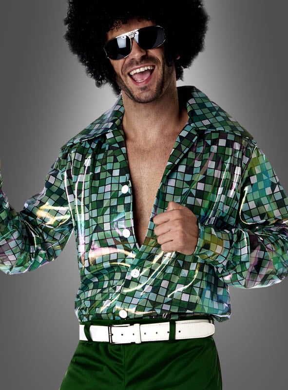 Ein Mann, der das „Disco Tino“-Outfit im Stil der 70er Jahre trägt – bestehend aus einem glänzenden, gemusterten Hemd, einer grünen Hose, einem weißen Gürtel, einer übergroßen Sonnenbrille und einer Afro-Perücke –, lächelt begeistert vor einem grauen Hintergrund.