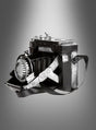 Eine Vintage-Fotokamera-Dekoration aus Blech mit Trageriemen, präsentiert vor einem grau abgestuften Hintergrund, eignet sich ideal als Vintage-Dekorationsstück oder als authentische Vintage-Fotokamera für jedes Kostüm-Ensemble im Stil der 20er Jahre.
