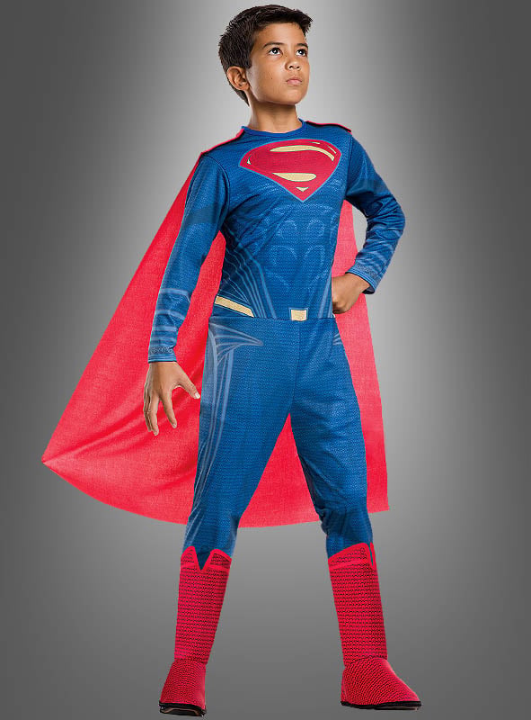 Ein kleiner Junge steht selbstbewusst in seinem Superman-Kinderkostüm der Justice League – einem blauen Anzug mit rotem Umhang und Stiefeln – vor einem schlichten grauen Hintergrund und ist bereit, sich dem Heldenteam anzuschließen.