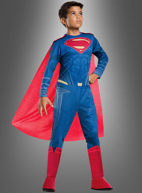 Ein kleiner Junge steht selbstbewusst in seinem Superman-Kinderkostüm der Justice League – einem blauen Anzug mit rotem Umhang und Stiefeln – vor einem schlichten grauen Hintergrund und ist bereit, sich dem Heldenteam anzuschließen.