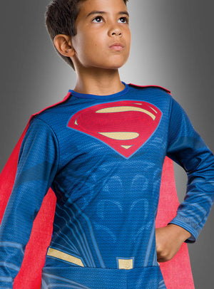 Ein kleiner Junge in einem Superman-Kinderkostüm der Justice League mit blauem Anzug und rotem Umhang steht mit der Hand in der Hüfte vor einem grauen Hintergrund und blickt nach oben – bereit, sich der Justice League anzuschließen.