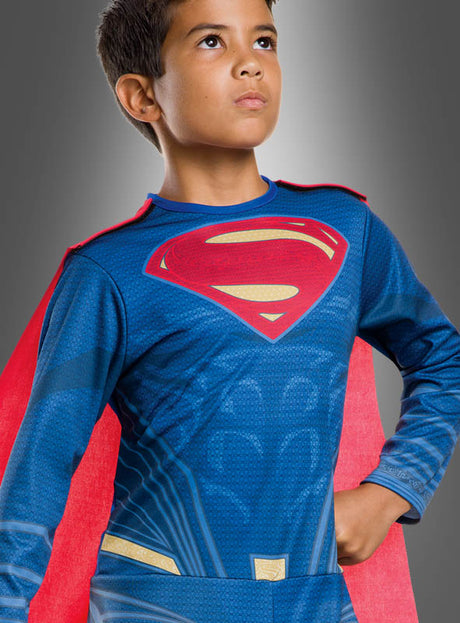 Ein kleiner Junge in einem Superman-Kinderkostüm der Justice League mit blauem Anzug und rotem Umhang steht mit der Hand in der Hüfte vor einem grauen Hintergrund und blickt nach oben – bereit, sich der Justice League anzuschließen.