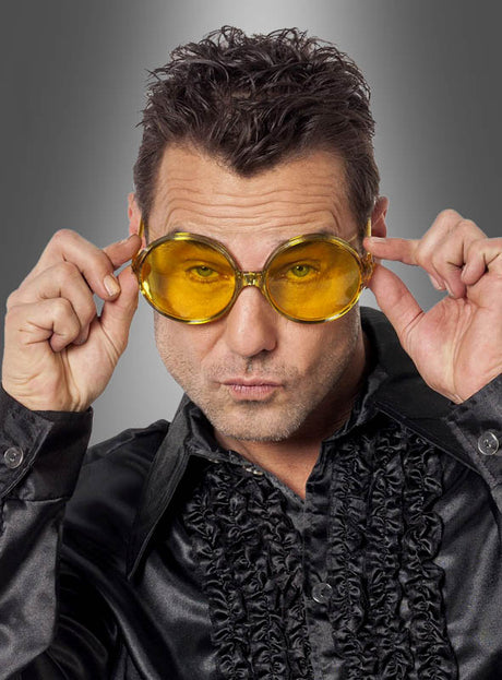 Ein Mann, der eine 70er-Partybrille mit großen Gläsern in leuchtendem Gelb und ein glänzendes schwarzes Rüschenhemd trägt, hält die übergroße Brille mit beiden Händen vor einem grauen Farbverlaufshintergrund – perfekt als Accessoire für eine Schlagerparty oder zur Vervollständigung deines Hippie-Kostüms.