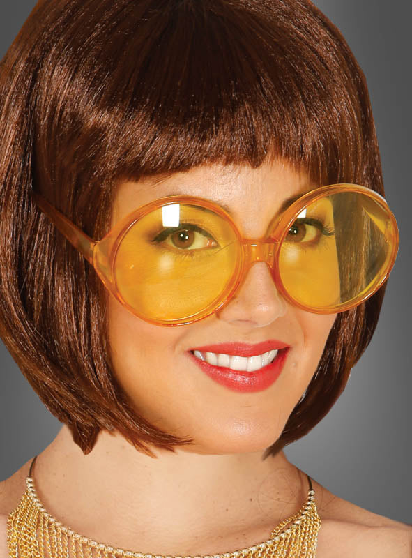 Lächelnde Frau mit kurzen braunen Haaren, die eine 70er-Jahre-Partybrille mit großen Gläsern und eine Halskette trägt und vor einem schlichten grauen Hintergrund posiert.