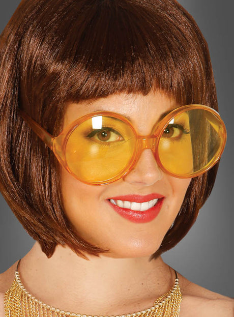 Lächelnde Frau mit kurzen braunen Haaren, die eine 70er-Jahre-Partybrille mit großen Gläsern und eine Halskette trägt und vor einem schlichten grauen Hintergrund posiert.