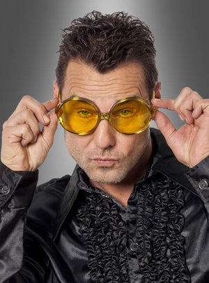 Ein Mann, der eine große gelbe 70er-Partybrille mit großen Gläsern und ein glänzendes schwarzes Rüschenhemd trägt, die Brille mit beiden Händen festhält und die Lippen spitzt, posiert vor einem grauen Hintergrund – ein perfektes Schlagerparty-Accessoire für jedes Hippie-Kostüm.