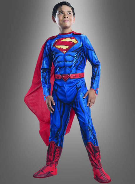 Ein kleiner Junge steht selbstbewusst in seinem Superman-Overall für Kinder, der in den klassischen Farben Blau und Rot gehalten ist und über einen leuchtend roten Umhang verfügt – perfekt für jede Comic-Helden-Geburtstagsfeier oder jede Fasching-Kostümparty – vor einem grauen Hintergrund mit Farbverlauf.
