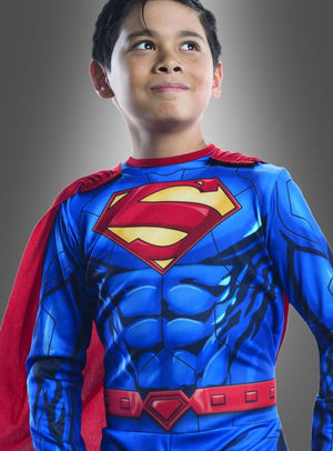 Ein Kind in einem blauen Superman-Overall für Kinder mit rotem Umhang steht vor einem schlichten grauen Hintergrund, blickt leicht nach oben und lächelt – die perfekte Kleidung für eine Comic-Helden-Geburtstagsfeier oder eine Faschingskostümparty.