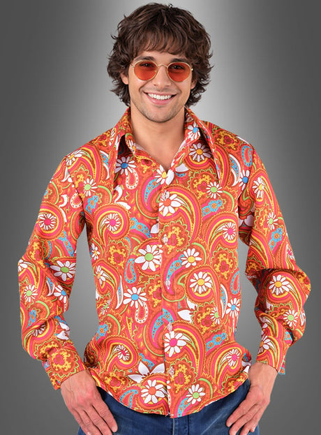 Ein Mann, der das „Schlagerparty-Hemd Martin für Herren“ mit einem bunten Paisley- und Blumenmuster trägt, dazu blaue Jeans und eine orangefarbene Sonnenbrille, steht vor einem grauen Hintergrund mit Farbverlauf.