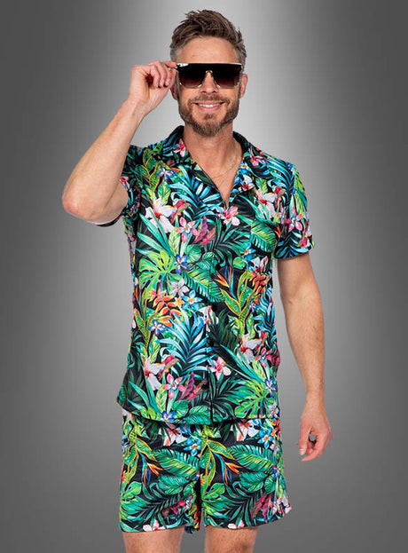 Ein Mann mit Sonnenbrille und einem farbenfrohen „Tropisches Hawaii“-Set aus Hemd und Shorts steht vor einem grauen Hintergrund und lächelt in die Kamera – und präsentiert damit das perfekte Sommeroutfit.