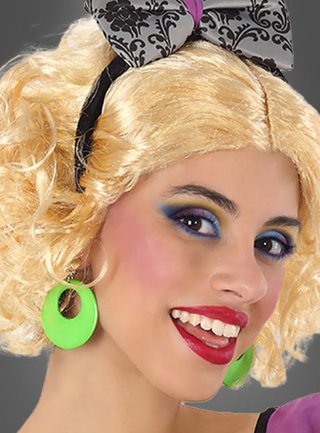 Die Person mit den blonden Locken lächelt in die Kamera und trägt leuchtend grüne Clip-Ohrringe im Retro-Look, buntes Make-up und eine gemusterte Haarschleife.