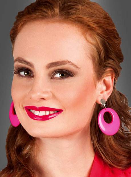 Eine Frau mit langen roten Haaren, die knallpinken Lippenstift, Ohrclips im Retro-Look und ein rosa Oberteil trägt, lächelt vor einem schlichten grauen Hintergrund.
