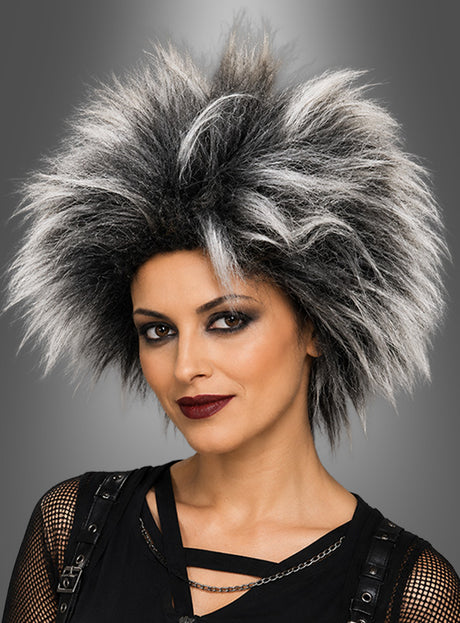 Ein Model präsentiert die Perücke „Punkerin – Igelfrisur“ mit dramatischer schwarz-weißer Stachelfrisur, auffälligem Augen-Make-up und dunklem Lippenstift. Es trägt ein schwarzes Oberteil mit Ketten vor einem grauen Hintergrund mit Farbverlauf.