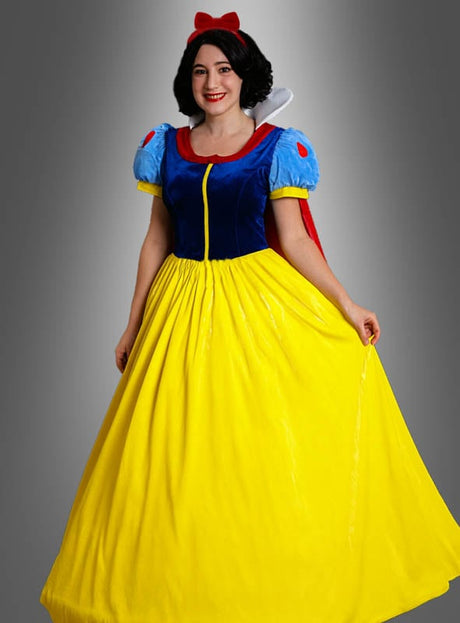 Eine Person, die das von Schneewittchen inspirierte „Märchenprinzessin Schneewittchen“-Kostüm trägt – bestehend aus einem blauen Oberteil, einem gelben Rock, einem roten Haarband mit Schleife und einem roten Umhang –, steht vor einem grauen Hintergrund.