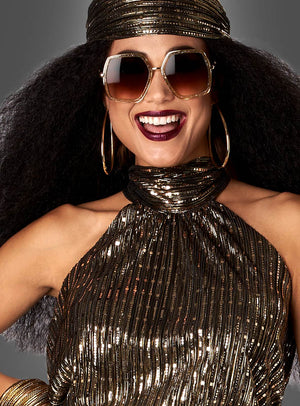 Eine Frau lächelt vor einem neutralen Hintergrund. Sie trägt den 70er-Jahre-Disco-Zweiteiler „Gold Fever“, dazu eine große Sonnenbrille und goldene Creolen.