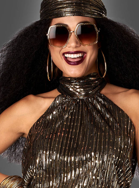 Eine Frau lächelt vor einem neutralen Hintergrund. Sie trägt den 70er-Jahre-Disco-Zweiteiler „Gold Fever“, dazu eine große Sonnenbrille und goldene Creolen.