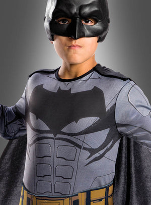Ein Kind trägt das Batman-Filmkostüm für Kinder aus „Justice League“ mit einer schwarzen Maske, einem aufgedruckten Fledermaus-Symbol auf der Brust und einem goldenen Werkzeuggürtel und steht vor einem grauen Hintergrund. Dieses Kostüm ist ein offizielles DC-Comics-Lizenzprodukt.