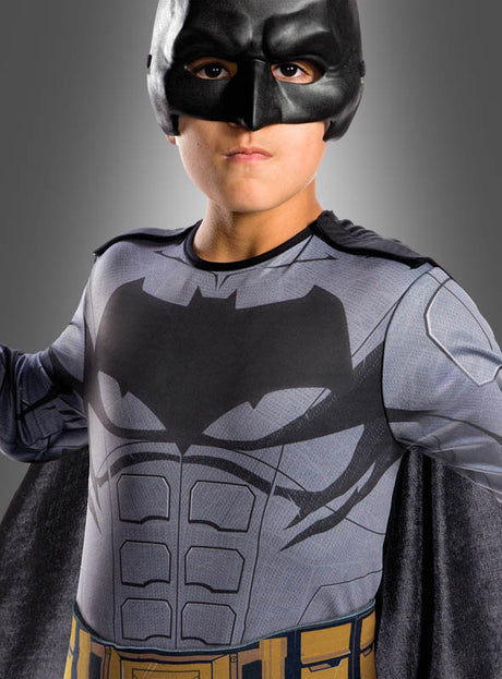Ein Kind trägt das Batman-Filmkostüm für Kinder aus „Justice League“ mit einer schwarzen Maske, einem aufgedruckten Fledermaus-Symbol auf der Brust und einem goldenen Werkzeuggürtel und steht vor einem grauen Hintergrund. Dieses Kostüm ist ein offizielles DC-Comics-Lizenzprodukt.