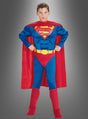 Ein Kind, das das Original-Superman-Kinderkostüm trägt – bestehend aus einem roten Umhang, einem blauen Anzug, roten Stiefeln und einem gelben Gürtel –, steht mit den Händen in den Hüften da.