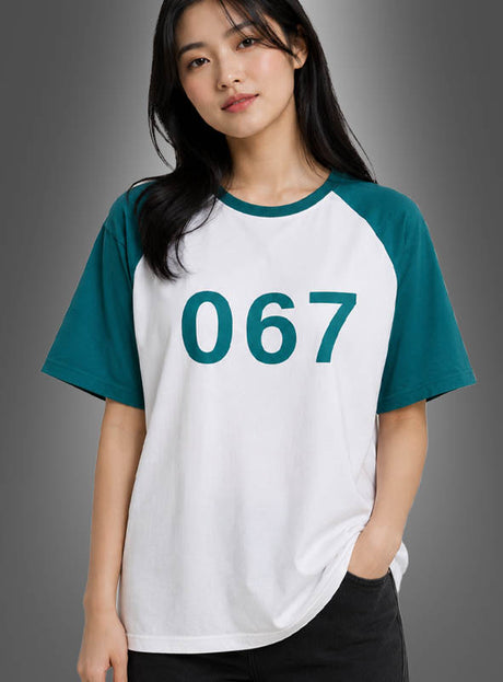 Vor einem schlichten grauen Hintergrund steht eine Frau, die das „Game Spieler Nummer 067“-T-Shirt für Damen trägt – ein weiß-grünes Raglan-Shirt mit der Nummer 067 auf der Vorderseite, das sich perfekt als Damenkostüm für das „Squad Game“-Cosplay eignet.