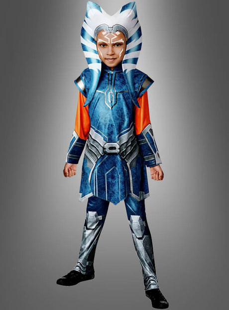 Ein Kind im „Ahsoka Tano Kostüm 2-Teiler Star Wars“ – bestehend aus einem blauen Outfit, einer gestreiften Kopfbedeckung und Gesichtsbemalung – steht vor einem grauen Hintergrund. Dieses detailreiche Kinderkostüm ist von Ahsokas kultigem Look aus der „Clone Wars“-Kostümkollektion inspiriert.