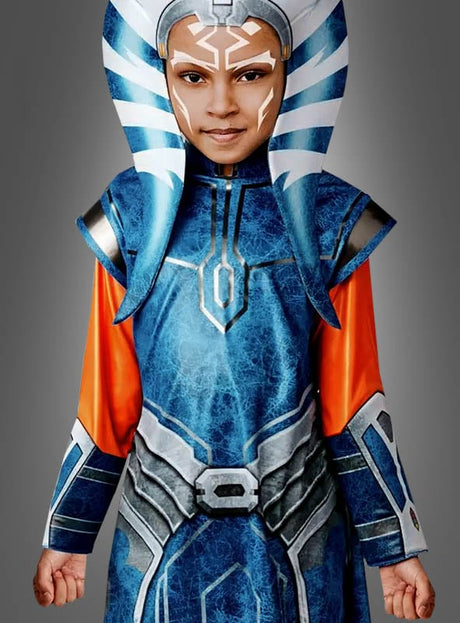 Ein Kind, das das „Ahsoka Tano Kostüm 2-Teiler Star Wars“ trägt – ein blaues, orangefarbenes und weißes Science-Fiction-Kostüm, das von „Clone Wars“ inspiriert ist und mit passendem Kopfschmuck sowie Gesichtsbemalung ergänzt wird –, steht vor einem schlichten grauen Hintergrund.