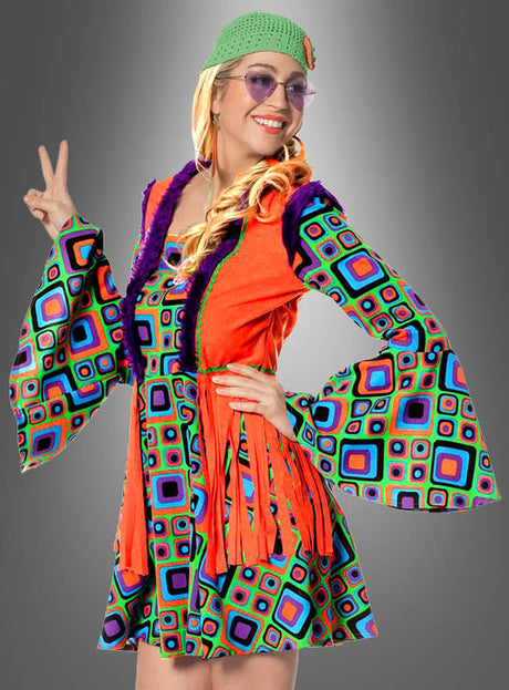 Eine Person in einem farbenfrohen Hippiekleid mit Fransenweste und geometrischen Mustern, einer grünen Mütze, einer getönten Brille, die vor einem grauen Hintergrund das Peace-Zeichen macht – ein echtes Blumenkind, perfekt gekleidet für jede 70er-Jahre-Kostümparty.