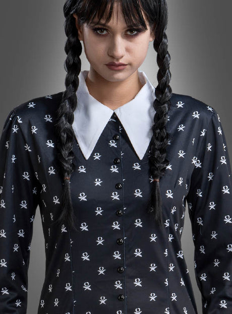 Eine Frau mit geflochtenen Haaren trägt das „Wednesday Damen Kleid schwarz-weiß“ – ein schwarzes Kleid mit weißen Totenkopfmustern und einem weißen spitzen Kragen –, das an das ikonische Wednesday-Kostüm aus der Addams Family angelehnt ist, und steht vor einem grauen Hintergrund.