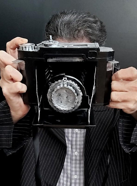 Eine Person in einem Nadelstreifenanzug hält eine große Vintage-Kamera aus Blech vor ihr Gesicht und richtet sie vor einem schlichten schwarzen Hintergrund auf den Betrachter – eine auffällige Anspielung auf den klassischen Stil und ein Kostümaccessoire der 20er Jahre.