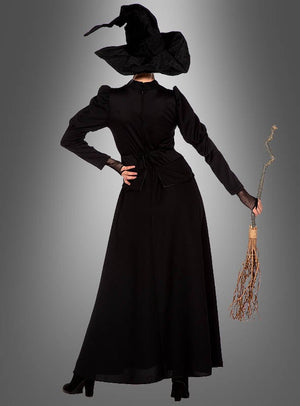 Eine Person trägt das „klassische schwarze Hexenkleid“ mit spitzem Hut, steht mit einem Besen in der Hand und dem Rücken zur Kamera vor einem grauen Hintergrund.