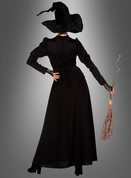 Eine Person trägt das „klassische schwarze Hexenkleid“ mit spitzem Hut, steht mit einem Besen in der Hand und dem Rücken zur Kamera vor einem grauen Hintergrund.