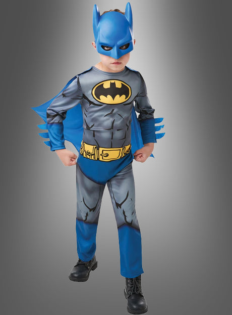 Ein Kind, das das Batman-Kinderkostüm trägt – bestehend aus einem blauen Umhang, einer Batman-Maske und einem gelben Gürtel –, steht mit geballten Fäusten vor einem grauen Hintergrund.