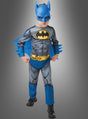 Ein Kind, das das Batman-Kinderkostüm trägt – bestehend aus einem blauen Umhang, einer Batman-Maske und einem gelben Gürtel –, steht mit geballten Fäusten vor einem grauen Hintergrund.