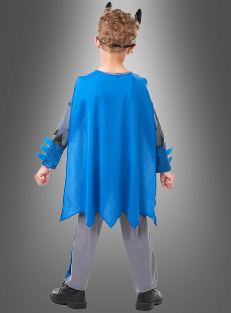 Ein Kind, das ein Batman-Kinderkostüm in den Farben Blau und Grau trägt, mit einem blauen Umhang und einem Stirnband mit schwarzen Ohren, steht mit dem Rücken zur Kamera vor einem grauen Hintergrund.