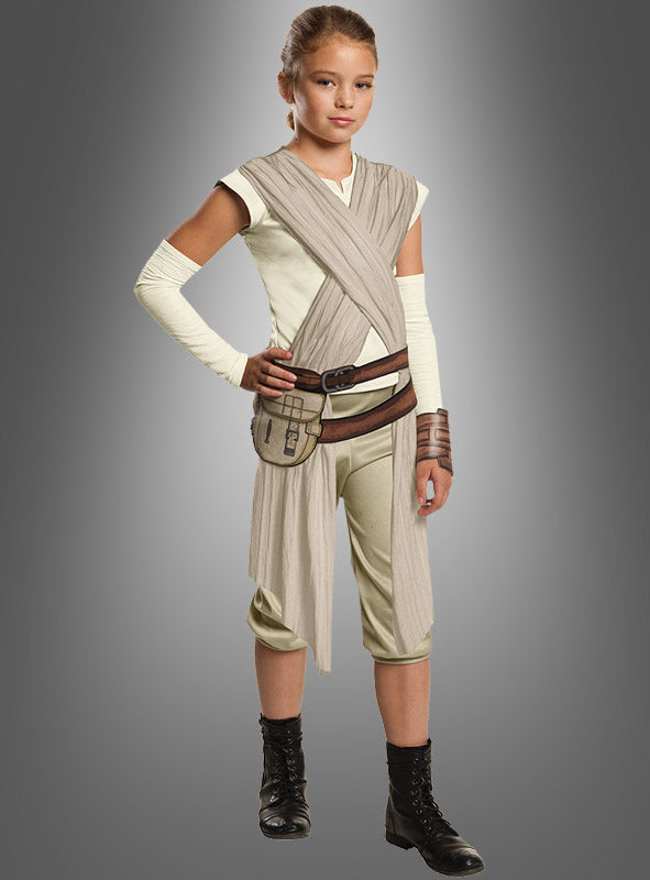 Vor einem grauen Hintergrund steht ein junges Mädchen, das das „Rey Star Wars Deluxe Kinderkostüm“ trägt, ein offizielles Lizenzkostüm mit beigen Wickeltüchern, einem braunen Gürtel, Armstulpen und schwarzen Stiefeln.