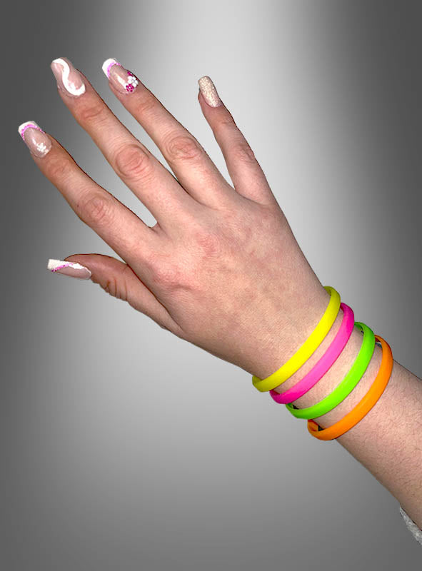 Eine Hand mit verzierten Fingernägeln trägt vier bunte Silikon-Neonarmbänder im 80er-Jahre-Stil – in Gelb, Pink, Grün und Orange –, die vor einem grauen Hintergrund mit Farbverlauf zur Geltung kommen und dieses lebhafte 4er-Set aus Silikon-Neonarmbändern im 80er-Jahre-Stil perfekt in Szene setzen.