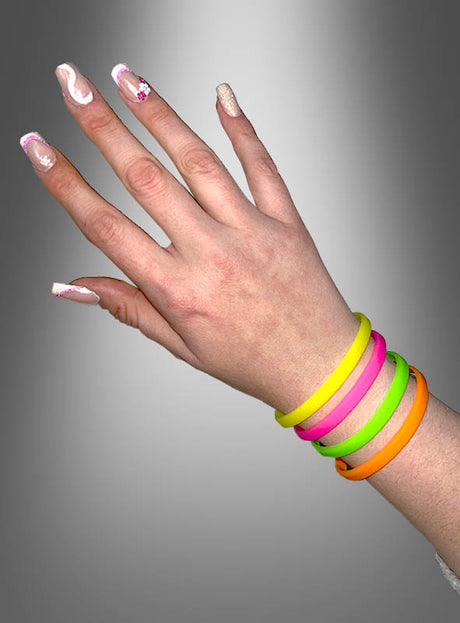 Eine Hand mit verzierten Fingernägeln trägt vier bunte Silikon-Neonarmbänder im 80er-Jahre-Stil – in Gelb, Pink, Grün und Orange –, die vor einem grauen Hintergrund mit Farbverlauf zur Geltung kommen und dieses lebhafte 4er-Set aus Silikon-Neonarmbändern im 80er-Jahre-Stil perfekt in Szene setzen.