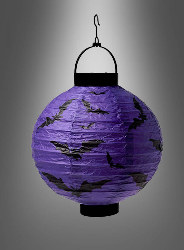 Die LED-Halloween-Papierlaterne „Fledermaus oder Spinnen“ – eine violette Laterne, die mit schwarzen Fledermaus-Silhouetten verziert ist und an einem schwarzen Haken hängt – leuchtet hell als leuchtender Lampion vor einem grauen Hintergrund mit Farbverlauf und ist damit die perfekte, auffällige Halloween-Dekoration.