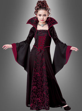 Ein junges Mädchen trägt das im Gothic-Stil gehaltene „Vampir-Königin“-Kinderkostüm „Mina“ mit hohem Kragen, ausgestellten Ärmeln und auffälligem rot-schwarz gemustertem Stoff und steht vor einem schlichten grauen Hintergrund. Dieses elegante Kostüm ist die ideale Wahl als Halloween-Kinderkostüm für alle, die den klassischen Vampir-Look lieben.