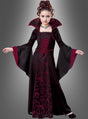 Ein junges Mädchen trägt das im Gothic-Stil gehaltene „Vampir-Königin“-Kinderkostüm „Mina“ mit hohem Kragen, ausgestellten Ärmeln und auffälligem rot-schwarz gemustertem Stoff und steht vor einem schlichten grauen Hintergrund. Dieses elegante Kostüm ist die ideale Wahl als Halloween-Kinderkostüm für alle, die den klassischen Vampir-Look lieben.