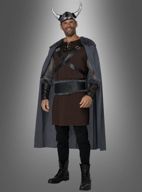 Ein Mann in einem Wikinger-Ragnar-Kostüm mit einem gehörnten Wikingerhelm, einem grauen Umhang, einer braunen Tunika, einer schwarzen Hose und schwarzen Stiefeln, der vor einem grauen Hintergrund mit Farbverlauf steht und lächelt.