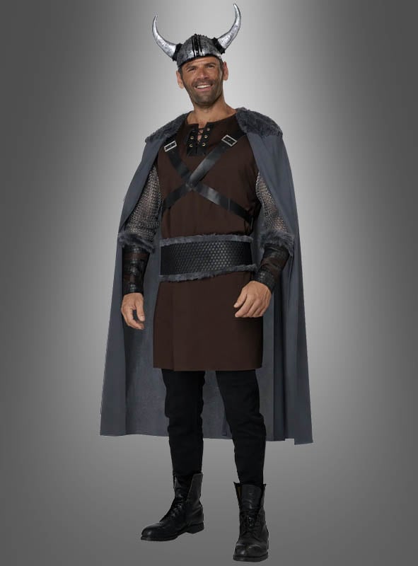 Ein Mann in einem Wikinger-Ragnar-Kostüm mit einem gehörnten Wikingerhelm, einem grauen Umhang, einer braunen Tunika, einer schwarzen Hose und schwarzen Stiefeln, der vor einem grauen Hintergrund mit Farbverlauf steht und lächelt.