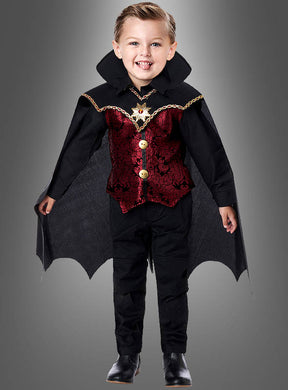 Ein kleines Kind trägt das „Edler Dracula“-Kostüm für Kinder mit auffälligen schwarzen und roten Details und einem wallenden Umhang und steht lächelnd vor einem schlichten grauen Hintergrund.