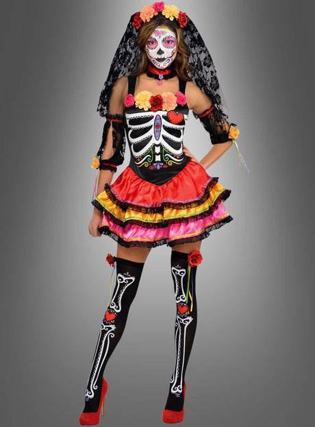 Vor einem grauen Hintergrund steht eine Frau, die das „Dia de los Muertos“-Komplettkostüm für Damen trägt, das sich durch farbenfrohe Skelettmotive, Sugar-Skull-Make-up, einen Spitzen-Schleier und rote Pumps auszeichnet.