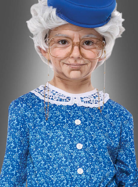 Ein Kind, das das „Oma-Kostüm für Mädchen“ trägt – bestehend aus einer grauen Perücke, einer goldgerahmten Brille, einem blauen Blumenkleid und einem dazu passenden blauen Hut –, posiert vor einem grauen Hintergrund.