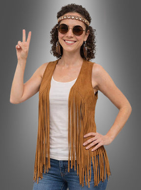 Eine Frau mit Sonnenbrille und Stirnband präsentiert die „Hippie Fransenweste mit Zubehör Damen“ über einem weißen Tanktop und Jeans und macht vor einem grauen Hintergrund das Peace-Zeichen.