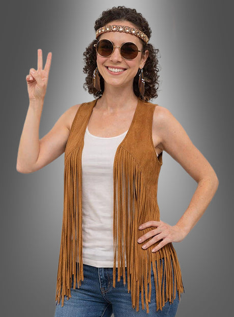 Eine Frau mit Sonnenbrille und Stirnband präsentiert die „Hippie Fransenweste mit Zubehör Damen“ über einem weißen Tanktop und Jeans und macht vor einem grauen Hintergrund das Peace-Zeichen.