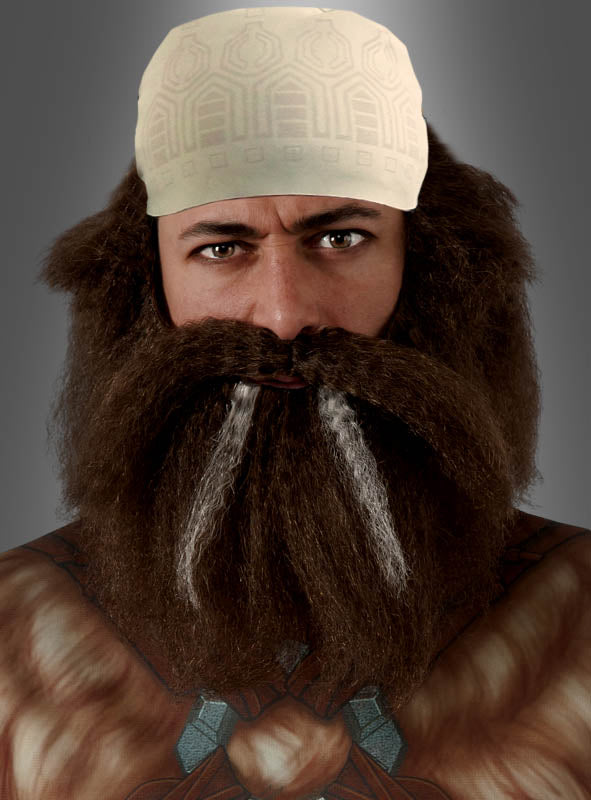 Eine Person, die das Dwalin-Bartset aus „Der Hobbit“ trägt, bestehend aus einem dichten braunen Bart mit weißen Strähnen, der an einen traditionellen Zwergenbart erinnert, kombiniert mit einer gemusterten cremefarbenen Kopfbedeckung und brauner Kleidung, die durch sich über Kreuzende blaue Riemen über der Brust akzentuiert wird.