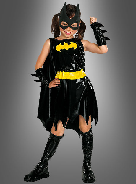 Ein Kind in einem „Batgirl Deluxe“-Kinderkostüm in Schwarz und Gelb posiert mit erhobenem Arm wie eine echte Superheldin. Vor einem grauen Hintergrund trägt sie eine Maske, einen Umhang, einen Gürtel, Armbänder und Stiefel.
